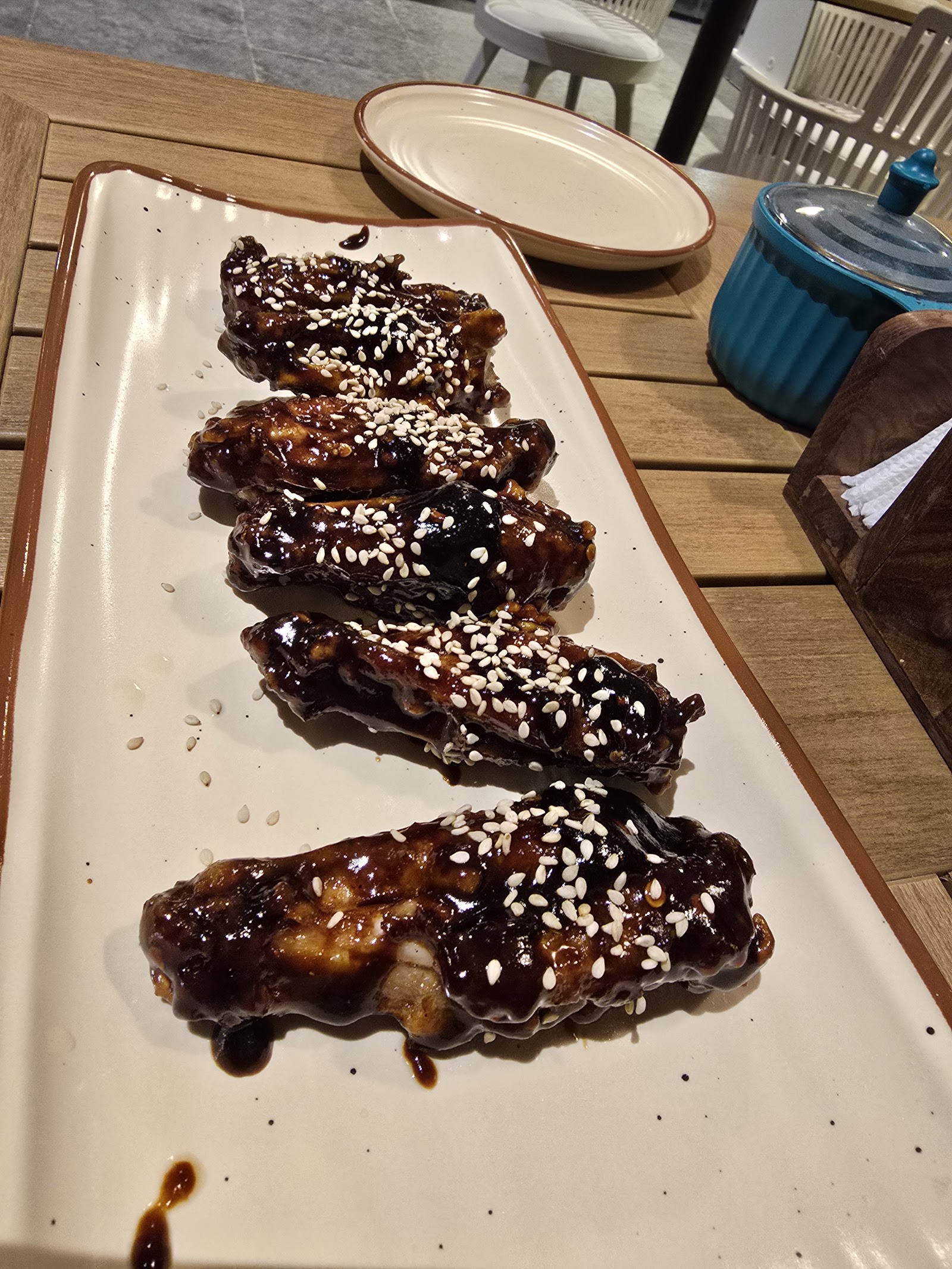 Five chicken wings in a glossy dark sauce with sesame seeds, on a long cream platter at a table in the cafe