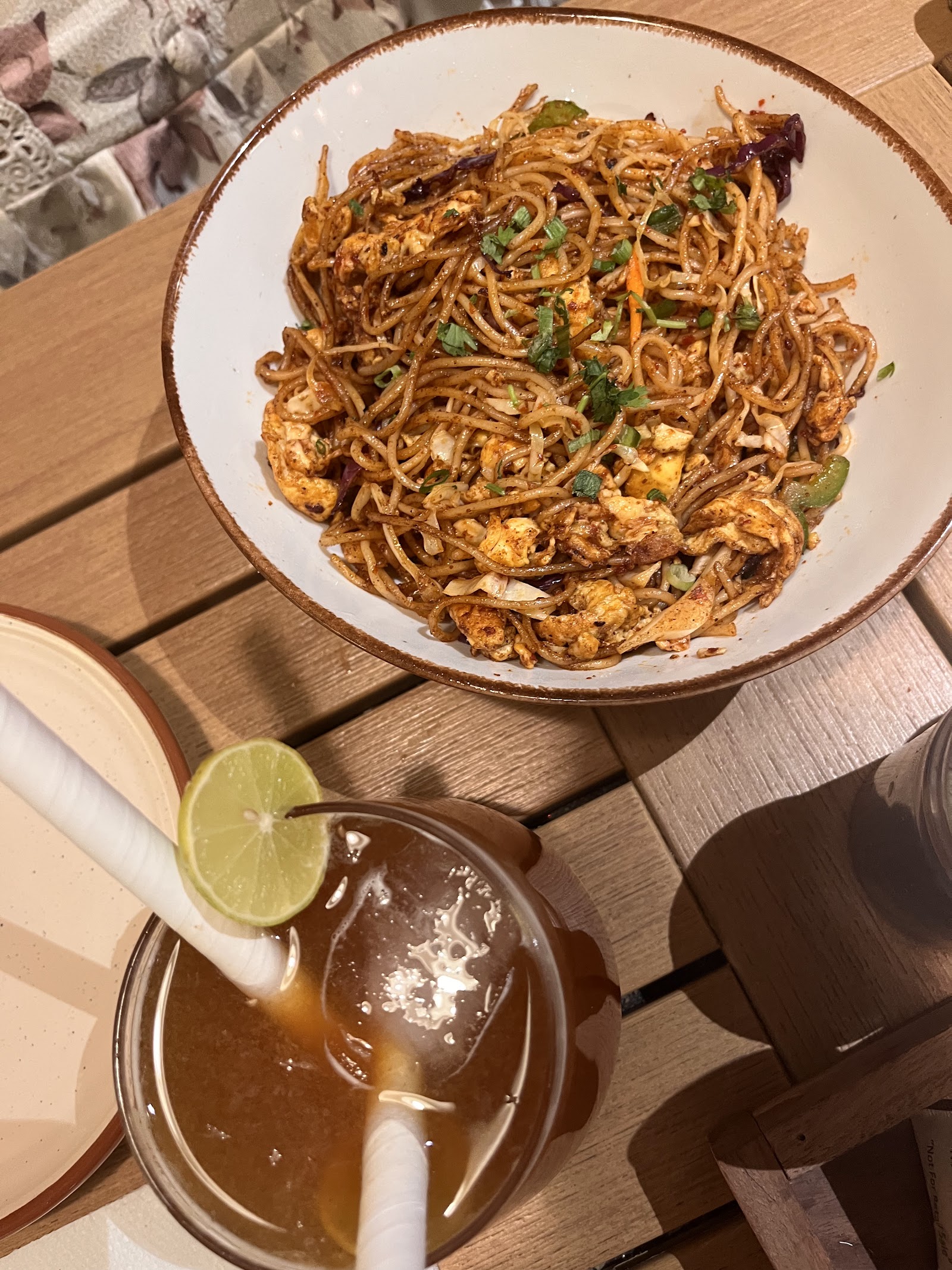 Chicken noodles in a stoneware bowl beside a tall iced tea with lime, on a wooden table