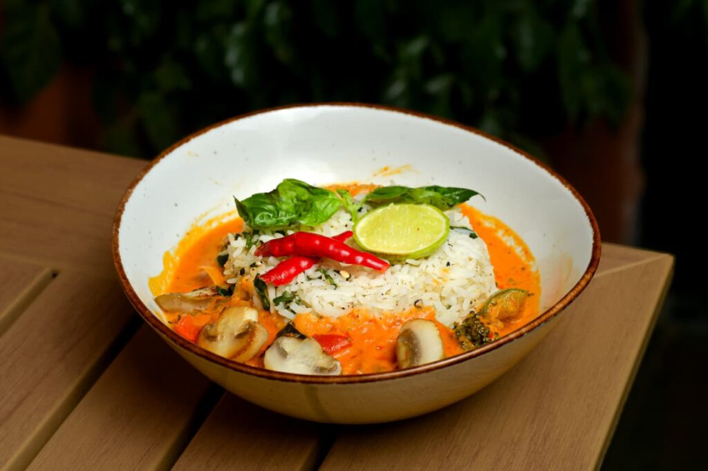 Thai green curry rice bowl in a stoneware bowl on a pale wooden table, with mushrooms, broccoli and a wedge of lime