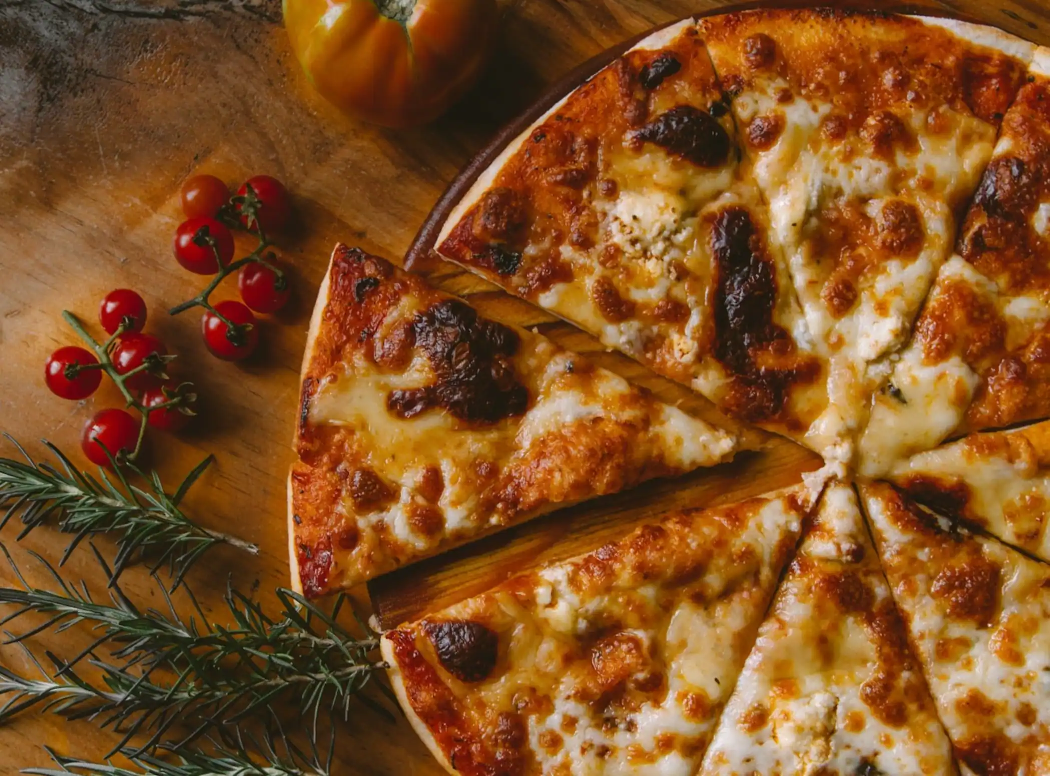 A stone-baked cheese pizza on a wooden board, cut into slices, with cherry tomatoes and rosemary alongside