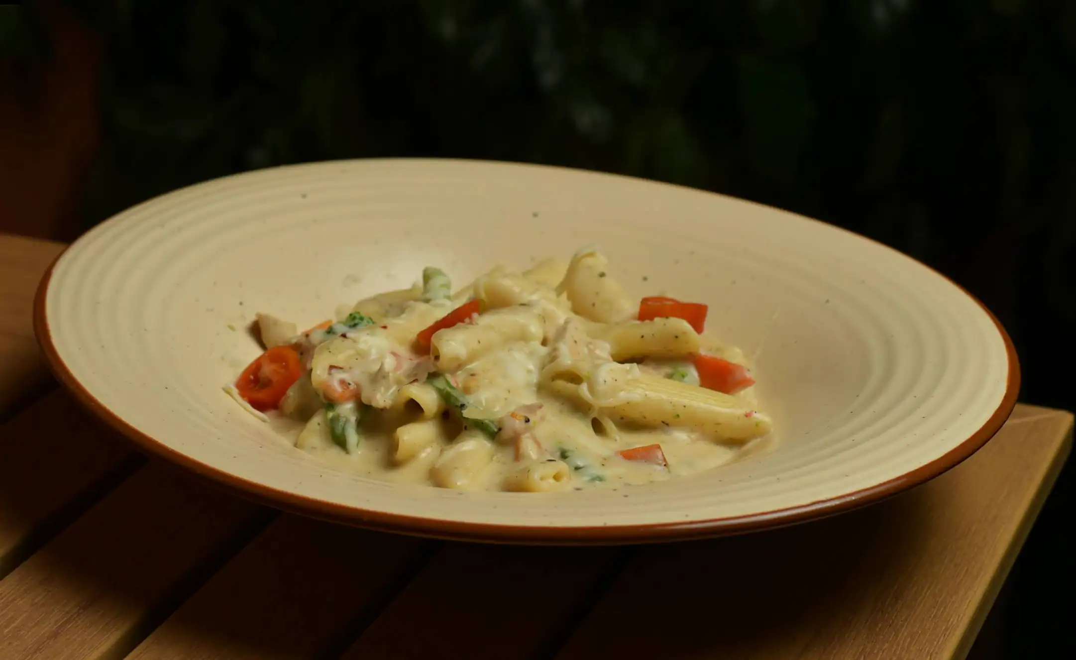 Alfredo penne in a cream bowl with red peppers and green beans, against a dark leafy background